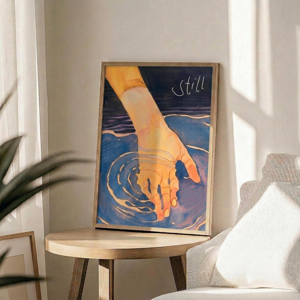 May include: Framed poster print displayed on round wooden side table next to beige upholstered chair. Artwork shows orange-toned hand reaching into dark blue water creating ripples, with "Still" text in white lettering against navy background. Natural lighting creates shadows across white wall. Green plant leaves visible in corner of minimalist room setting.