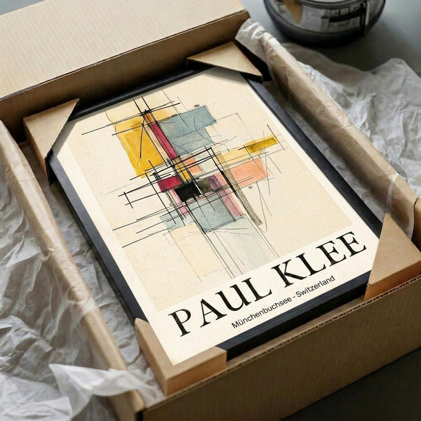 May include: Framed abstract art print with geometric shapes in yellow, red, blue, and pink watercolors intersected by black lines, displayed in black frame being unpacked from cardboard shipping box with protective wrapping paper on white surface