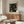 May include: Framed poster with black background featuring golden yellow dotted circles and cosmic patterns, displayed above wooden sideboard with sliding doors. Mid-century modern living room setting with cream upholstered swivel chair, glass-top oval coffee table, and white table lamp. Poster shows "YAYOI KUSAMA" and "Modern Art 1954" text with polka dot astronomical design reflecting on glossy table surface.