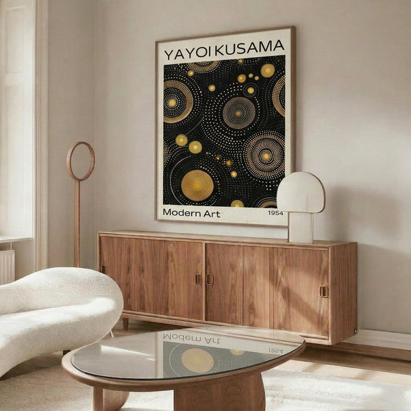 May include: Framed poster with black background featuring golden yellow dotted circles and cosmic patterns, displayed above wooden sideboard with sliding doors. Mid-century modern living room setting with cream upholstered swivel chair, glass-top oval coffee table, and white table lamp. Poster shows "YAYOI KUSAMA" and "Modern Art 1954" text with polka dot astronomical design reflecting on glossy table surface.
