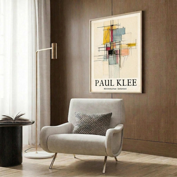 May include: Abstract geometric artwork poster with colorful overlapping rectangles in yellow, red, blue, and orange connected by black lines, displayed in light wood frame on textured wood paneled wall. Modern beige armchair with geometric patterned pillow sits below artwork, alongside brass floor lamp and dark round side table with open book.
