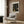 May include: Framed poster with black background and gold circular dot patterns hanging on wood-paneled wall above beige upholstered chair with geometric pillow. Modern brass floor lamp and round black side table with open book visible. Warm neutral tones throughout minimalist interior setting.