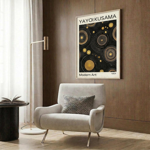May include: Framed poster with black background and gold circular dot patterns hanging on wood-paneled wall above beige upholstered chair with geometric pillow. Modern brass floor lamp and round black side table with open book visible. Warm neutral tones throughout minimalist interior setting.