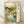May include: Framed Japanese woodblock-style poster print leaning against white wall on wooden floor. Artwork shows serene mountain landscape with winding stream, cherry blossoms, rocks, and distant sun in soft greens, blues, and peachy tones. Large vertical Japanese text on right side with smaller text below. Natural wood frame with light finish.
