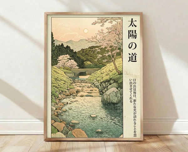 May include: Framed Japanese woodblock-style poster print leaning against white wall on wooden floor. Artwork shows serene mountain landscape with winding stream, cherry blossoms, rocks, and distant sun in soft greens, blues, and peachy tones. Large vertical Japanese text on right side with smaller text below. Natural wood frame with light finish.