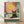 May include: Framed poster print leaning against white wall on light wood floor. Artwork shows black cat wearing round yellow sunglasses sitting poolside with turquoise water, coral-pink drink with striped straw and lemon slice nearby. Large yellow sun in coral-pink sky background. Retro-style illustration with warm coral, teal, and yellow color palette in natural wood frame.