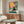 May include: Framed poster displayed above wooden sideboard in modern living room showing black cat wearing yellow sunglasses sitting by turquoise pool with pink cocktail, bright yellow sun in coral and teal tiled background. White curved chair and glass coffee table visible in foreground with neutral walls and contemporary decor.