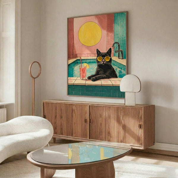 May include: Framed poster displayed above wooden sideboard in modern living room showing black cat wearing yellow sunglasses sitting by turquoise pool with pink cocktail, bright yellow sun in coral and teal tiled background. White curved chair and glass coffee table visible in foreground with neutral walls and contemporary decor.