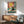 May include: Framed poster print hanging above wooden credenza showing black cat wearing yellow sunglasses lounging by turquoise swimming pool with coral drink nearby, bright yellow sun in coral sky background, displayed in modern living room with mint green mid-century chair and white ceramic lamp