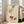 May include: Vintage-style art print in light wood frame leaning against white wall next to beige sofa. Artwork shows hand dropping jewelry into orange bowl, with French text below on cream aged paper background. Natural lighting from windows creates soft shadows on wooden floor.