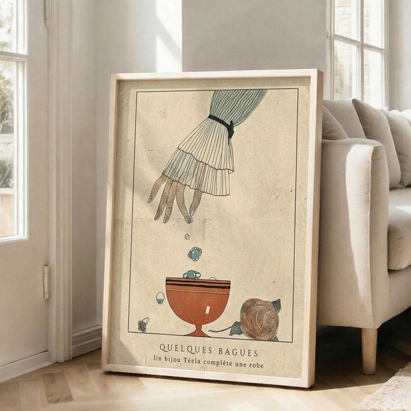 May include: Vintage-style art print in light wood frame leaning against white wall next to beige sofa. Artwork shows hand dropping jewelry into orange bowl, with French text below on cream aged paper background. Natural lighting from windows creates soft shadows on wooden floor.