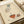 May include: Vintage-style Art Deco poster print displayed in an open book or portfolio with cream-colored pages and beige ribbon binding. The illustration features a woman's lower body in a striped dress and stockings reaching toward an ornate orange teacup, with decorative swirls and geometric elements. The artwork has muted teal, orange, and brown colors on aged paper texture with French text at the bottom.