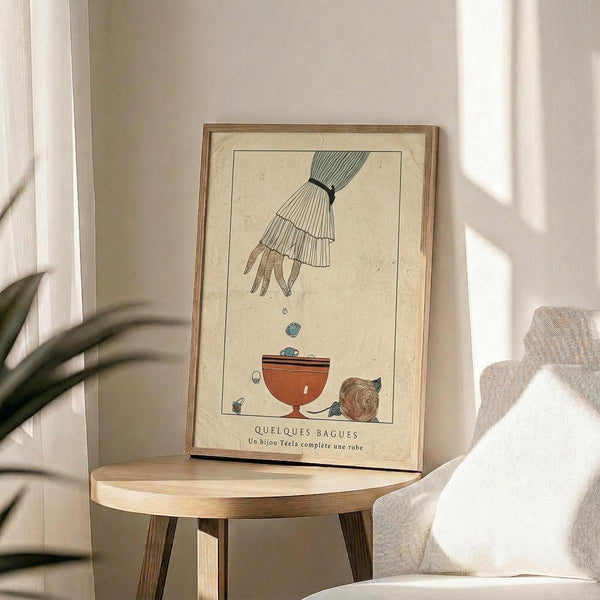 May include: Vintage Art Deco style poster print in wooden frame displayed on round wooden side table. Artwork shows elegant hand with flowing sleeve dropping rings into decorative bowl, rendered in muted tones of beige, green and terracotta. French text below illustration. Room setting features beige walls, white curtains with natural lighting, cream upholstered chair, and green plant leaves visible in corner.