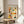 May include: Framed poster with twelve illustrated portrait panels showing diverse people in colorful clothing and headwear, displayed on a round wooden side table next to a beige upholstered chair. Natural sunlight streams across white walls, with green plant leaves visible in corner. The artwork features hand-drawn style figures in warm earth tones including orange, yellow, green, and blue, arranged in a grid format with "SOIS UNIQUE" text at top.