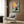 May include: Framed poster print featuring a black cat wearing yellow sunglasses lounging by a turquoise swimming pool with a tropical drink, yellow sun and coral-pink sky in background. Modern living room setting with beige armchair, geometric pillow, brass floor lamp, and wood-paneled wall. Warm natural lighting through window curtains.
