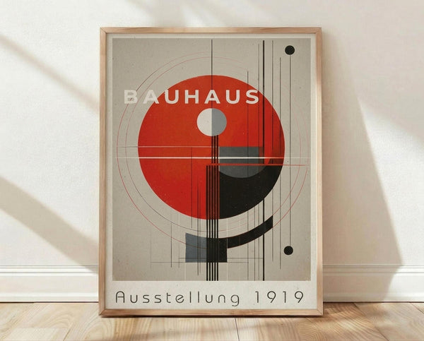 May include: Framed Bauhaus-style poster with geometric design featuring large red circle, black curved elements, and grid lines on beige background. The artwork displays "BAUHAUS" text and "Ausstellung 1919" at bottom. Light wood frame sits on wooden floor against white wall with natural shadow lighting.