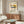 May include: Mid-century modern living room mockup showing cream-colored poster in thin frame above walnut credenza. Poster features colorful cantilever chair illustration with woven cane back, blue and red cushions, yellow circle background, and "1986 DESIGN REVOLUTION" text. White sculptural chair and glass-top coffee table in foreground, warm natural lighting creates shadows across wood surfaces.
