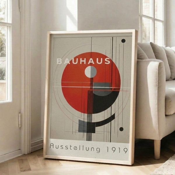 May include: Framed geometric art print in light wood frame leaning against white wall next to beige upholstered chair. Poster features red and black circular design with intersecting lines and geometric elements on cream background, with "BAUHAUS" and "Ausstellung 1919" text. Modern minimalist room setting with hardwood floors and natural lighting from windows.