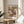 May include: Framed art print displaying colorful water lilies in pink, purple, yellow and white with green lily pads on dark background, positioned on round wooden side table next to beige upholstered chair. Natural sunlight streams across neutral wall creating soft shadows. Plant leaves visible in corner of bright, minimalist living room setting.