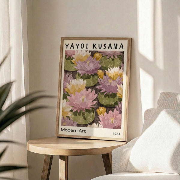 May include: Framed art print displaying colorful water lilies in pink, purple, yellow and white with green lily pads on dark background, positioned on round wooden side table next to beige upholstered chair. Natural sunlight streams across neutral wall creating soft shadows. Plant leaves visible in corner of bright, minimalist living room setting.