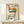 May include: Framed vintage-style poster featuring a modernist cantilever chair design with colorful geometric shapes - yellow circle, blue oval, woven cane back, and black tubular frame. Text reads "1986 DESIGN REVOLUTION" on cream background. Light wood frame displayed on minimalist wooden shelf with small white wrapped gift and pine sprig accent against neutral wall.