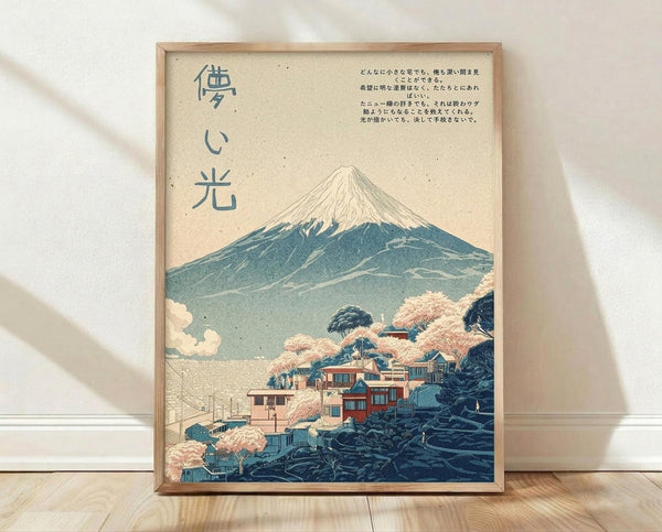 May include: Framed Japanese-style poster print leaning against white wall on light wooden floor. Artwork shows Mount Fuji with snow-capped peak rising above traditional village with pink cherry blossoms, rendered in vintage ukiyo-e woodblock print style using muted blues, pinks and cream tones. Features vertical Japanese text on left side and horizontal text on right. Natural wood frame with warm lighting casting soft shadows.