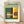 May include: Framed Bauhaus poster with modernist architectural buildings in geometric style, featuring green background with cream, orange, and yellow building facades. The vintage-inspired print shows "BAUHAUS" and "Ausstellung 1919" text. Light wood frame sits on wooden floor against white wall with natural lighting casting soft shadows.