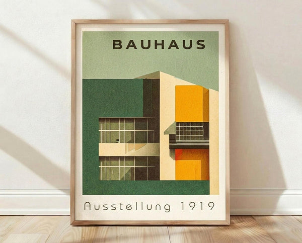 May include: Framed Bauhaus poster with modernist architectural buildings in geometric style, featuring green background with cream, orange, and yellow building facades. The vintage-inspired print shows "BAUHAUS" and "Ausstellung 1919" text. Light wood frame sits on wooden floor against white wall with natural lighting casting soft shadows.