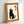 Framed artwork showing a black cat silhouette sitting upright with white paws and facial features, looking upward. Two Japanese kanji characters appear on the right side. The print has a cream-colored background with brown cork-style matting and light wooden frame. The piece leans against a white wall, casting a soft shadow.