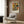 May include: Abstract art poster with geometric shapes in orange, yellow, green, and red tones displayed in white frame on wood-paneled wall. Modern reading corner features cream upholstered armchair with geometric pillow, brass floor lamp, and dark round side table with open book. Warm neutral interior with curtained window.