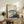 May include: Framed Japanese-style poster featuring Mount Fuji with snow-capped peak, traditional village buildings with pink cherry blossoms in foreground, and Japanese text characters. Print displayed on rustic wooden sideboard alongside fern plant, ceramic vases, and books in warm-toned living room with beige chair visible.
