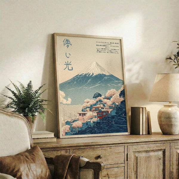 May include: Framed Japanese-style poster featuring Mount Fuji with snow-capped peak, traditional village buildings with pink cherry blossoms in foreground, and Japanese text characters. Print displayed on rustic wooden sideboard alongside fern plant, ceramic vases, and books in warm-toned living room with beige chair visible.