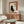 May include: Framed poster featuring black cat silhouette with Japanese characters on cream background with orange border, displayed above mid-century wooden credenza in modern living room with white curved chair and glass coffee table