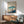 May include: Japanese-style poster print featuring Mount Fuji with snow-capped peak, traditional village buildings with pink cherry blossoms, and vertical Japanese text, displayed in thin wooden frame above mid-century modern walnut credenza with ceramic table lamp, next to sage green upholstered armchair in minimalist room with white walls and natural sunlight shadows