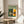 May include: Framed Bauhaus poster with geometric architectural building illustration in green, yellow, and orange colors, displayed on round wooden side table next to beige upholstered chair. Natural sunlight creates soft shadows on white wall behind the vintage-style exhibition print from 1919.