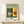 May include: Framed Bauhaus poster in light wood frame leaning against white wall on wooden shelf. Print features modernist building illustration in green, orange, and cream colors with geometric architecture and grid windows. Text reads "BAUHAUS" and "Ausstellung 1919" below the building design. Small white wrapped gift with greenery sits beside frame on light wood surface.
