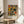 May include: Framed abstract art print with colorful geometric shapes in yellow, orange, green, and black displayed on a light wood round side table next to a beige upholstered chair. Natural sunlight streams across white walls creating soft shadows, with green plant leaves visible in the corner of the minimalist room setting.