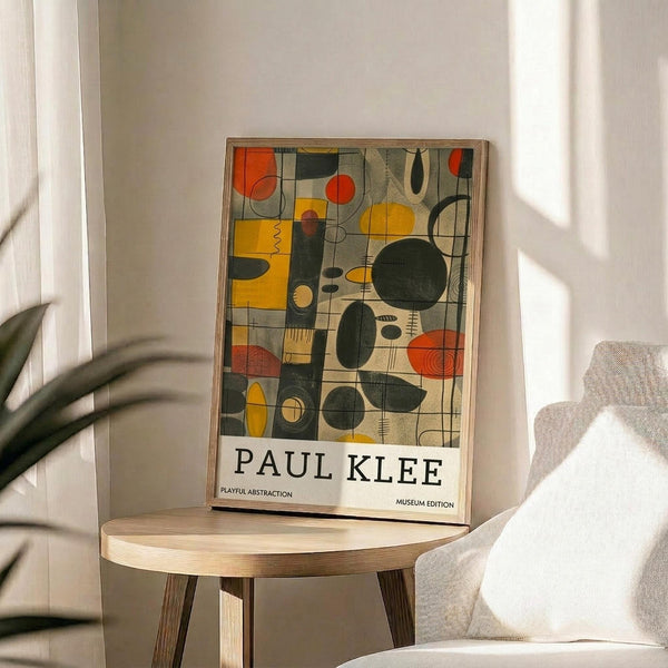 May include: Framed abstract art print with colorful geometric shapes in yellow, orange, green, and black displayed on a light wood round side table next to a beige upholstered chair. Natural sunlight streams across white walls creating soft shadows, with green plant leaves visible in the corner of the minimalist room setting.