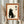 May include: Person holding framed artwork featuring black cat silhouette sitting upright with Japanese characters, displayed against cream background with orange border in light wood frame. Dried wheat stems visible on left side, soft natural lighting from window creates gentle shadows on neutral wall.