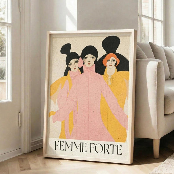May include: A large framed poster leaning against a beige sofa in a bright living room with white walls and windows. The artwork features three stylized women in Art Deco style with black hair and dramatic makeup, wearing flowing pink and yellow robes. The text "FEMME FORTE" appears at the bottom in black serif lettering. The poster has a cream background with a light wood frame and sits on hardwood flooring.