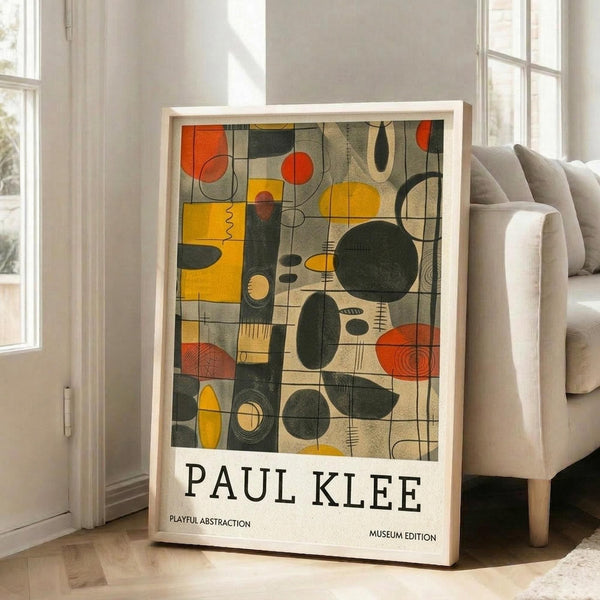 May include: A framed abstract art print leaning against a white wall next to a beige sectional sofa in a bright living room with large windows. The artwork features geometric shapes in yellow, orange, green, and black with playful lines and circles in mid-century modern style. Text reads "PAUL KLEE" and "PLAYFUL ABSTRACTION MUSEUM EDITION" below the colorful composition. Natural light streams across hardwood floors.