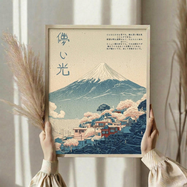 May include: Person holding framed Japanese-style artwork featuring Mount Fuji with snow-capped peak, traditional village buildings with cherry blossoms in foreground, and Japanese text. Art has vintage beige background with soft blue and pink tones. Natural wood frame, person wearing cream ruffled sleeves, dried pampas grass visible, soft window lighting in background.