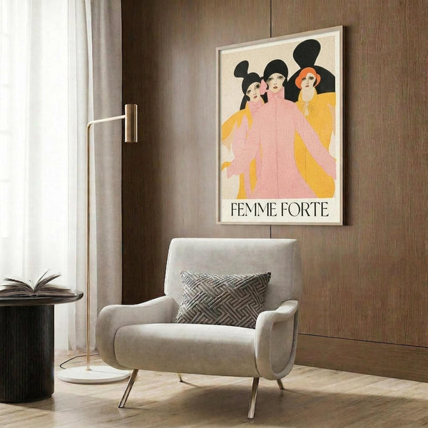 May include: Framed poster featuring three stylized women in pink and yellow clothing against black silhouettes with "FEMME FORTE" text below, displayed above a beige upholstered armchair with geometric patterned pillow in modern living room with wood paneled walls, gold floor lamp, and dark round side table with open book.