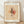 May include: Framed poster print leaning against white textured wall on light wood floor. Abstract modernist artwork features geometric shapes, lines, and watercolor-like washes in beige, pink, and black tones on aged paper background. Natural wood frame with white matting. Soft shadows cast across scene from natural lighting.
