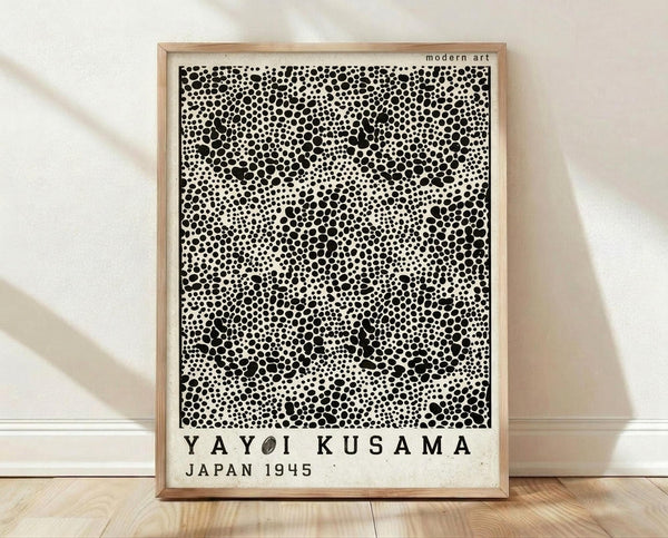 May include: Framed poster with black polka dot pattern on white background, text reading "YAYOI KUSAMA JAPAN 1945" below the artwork. Light wood frame with "modern art" text at top. Poster displayed on light wooden floor against white textured wall with soft natural lighting and shadows.