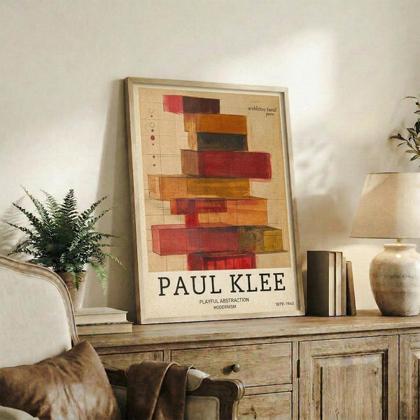 May include: Framed Paul Klee art print displayed on rustic wooden sideboard in warm living room setting. Abstract geometric composition features stacked colorful rectangular blocks in reds, oranges, yellows and browns on cream background with grid lines. Styled with fern plant, pottery vase, books, and brown leather chair visible. Natural lighting creates cozy atmosphere.