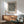 May include: Black and white dotted pattern poster in wooden frame hanging above mid-century modern wooden credenza with white ceramic lamp. Sage green upholstered armchair with wooden arms sits nearby on wooden floor. Bright natural light creates shadows on white wall in minimalist living room setting.