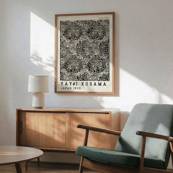 May include: Black and white dotted pattern poster in wooden frame hanging above mid-century modern wooden credenza with white ceramic lamp. Sage green upholstered armchair with wooden arms sits nearby on wooden floor. Bright natural light creates shadows on white wall in minimalist living room setting.
