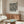 May include: Black and white dotted pattern poster with Japanese artist name and "Japan 1945" text, displayed above wooden sideboard in modern living room setting. White curved chair and glass-top coffee table visible. Poster shows polka dot artwork in thin frame against beige wall.
