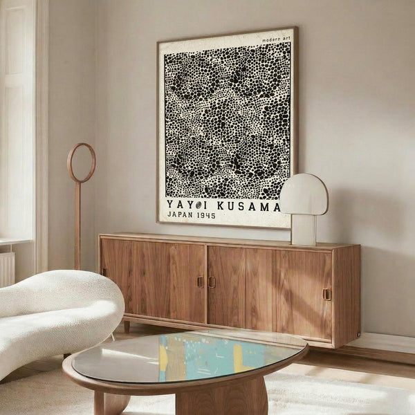 May include: Black and white dotted pattern poster with Japanese artist name and "Japan 1945" text, displayed above wooden sideboard in modern living room setting. White curved chair and glass-top coffee table visible. Poster shows polka dot artwork in thin frame against beige wall.